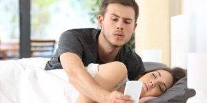 Man looking at sleeping wife's phone, considering the installation of spyware